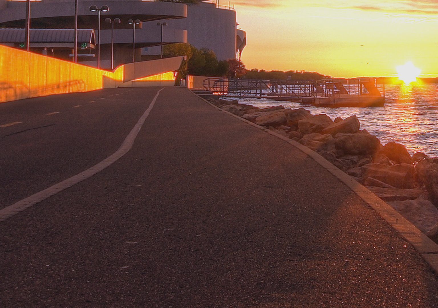 Paved bike path on lakeshore in the foreground with sun rising over horizon in the distance