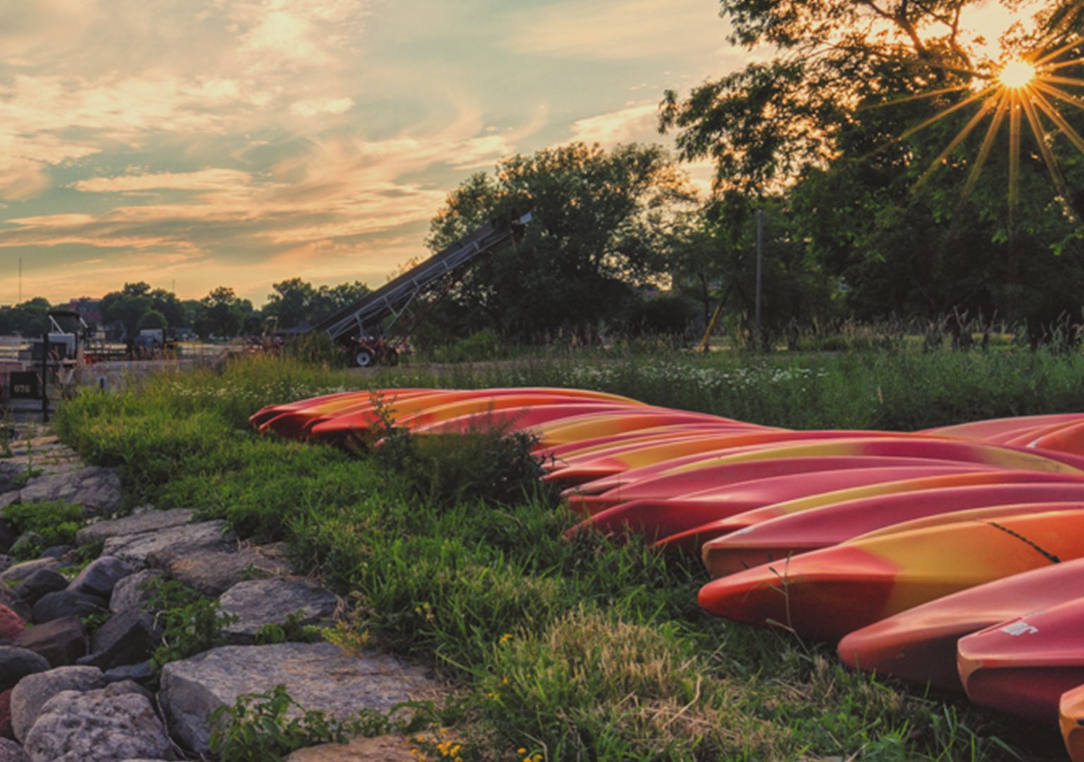 Canoes line the shore of Lake Mendota as the sun sets behind grove of trees in background