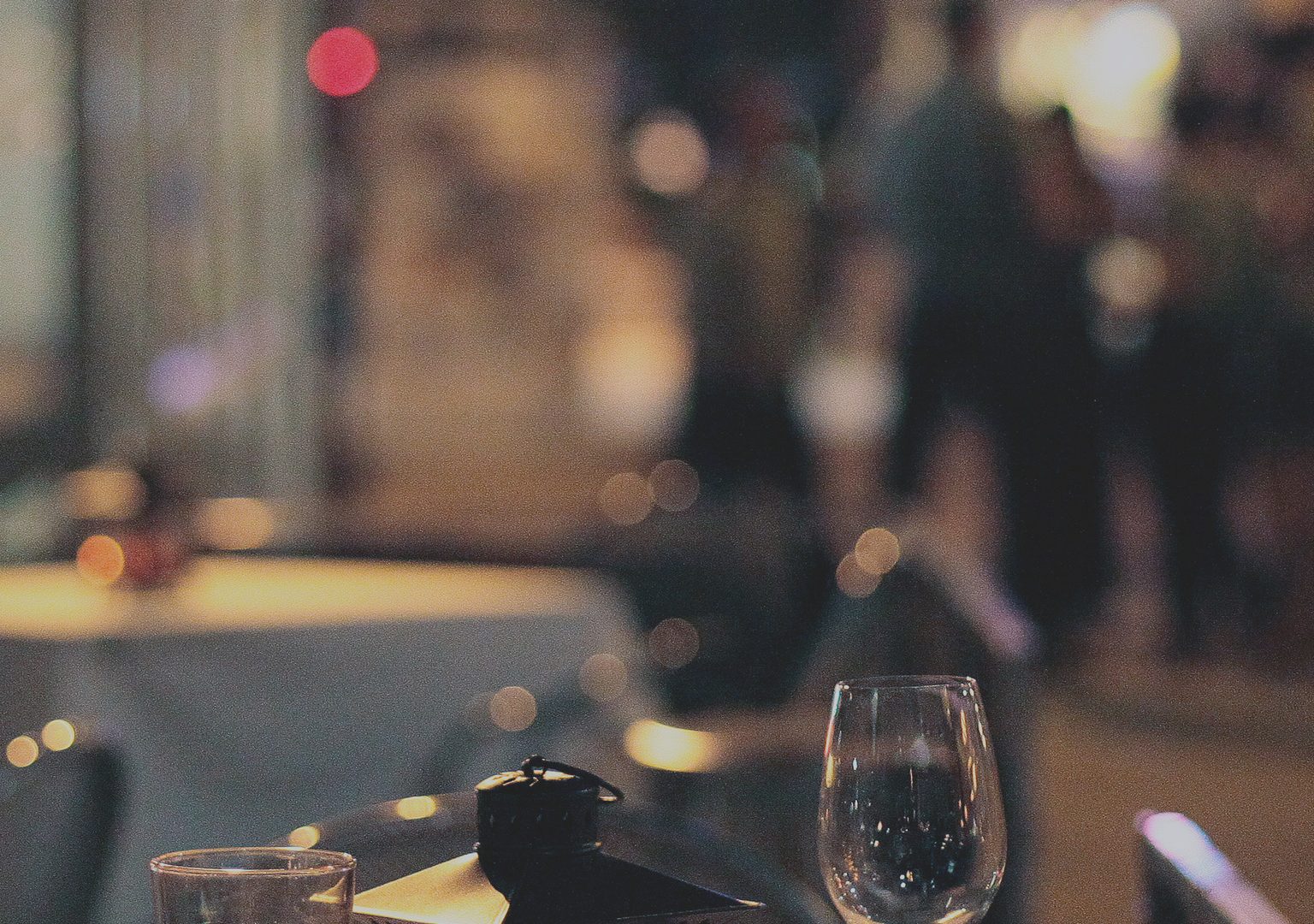 Restaurant table outdoors at nighttime with lantern and wine and water glasses in foreground, bus stop and people on sidewalk can be seen in background